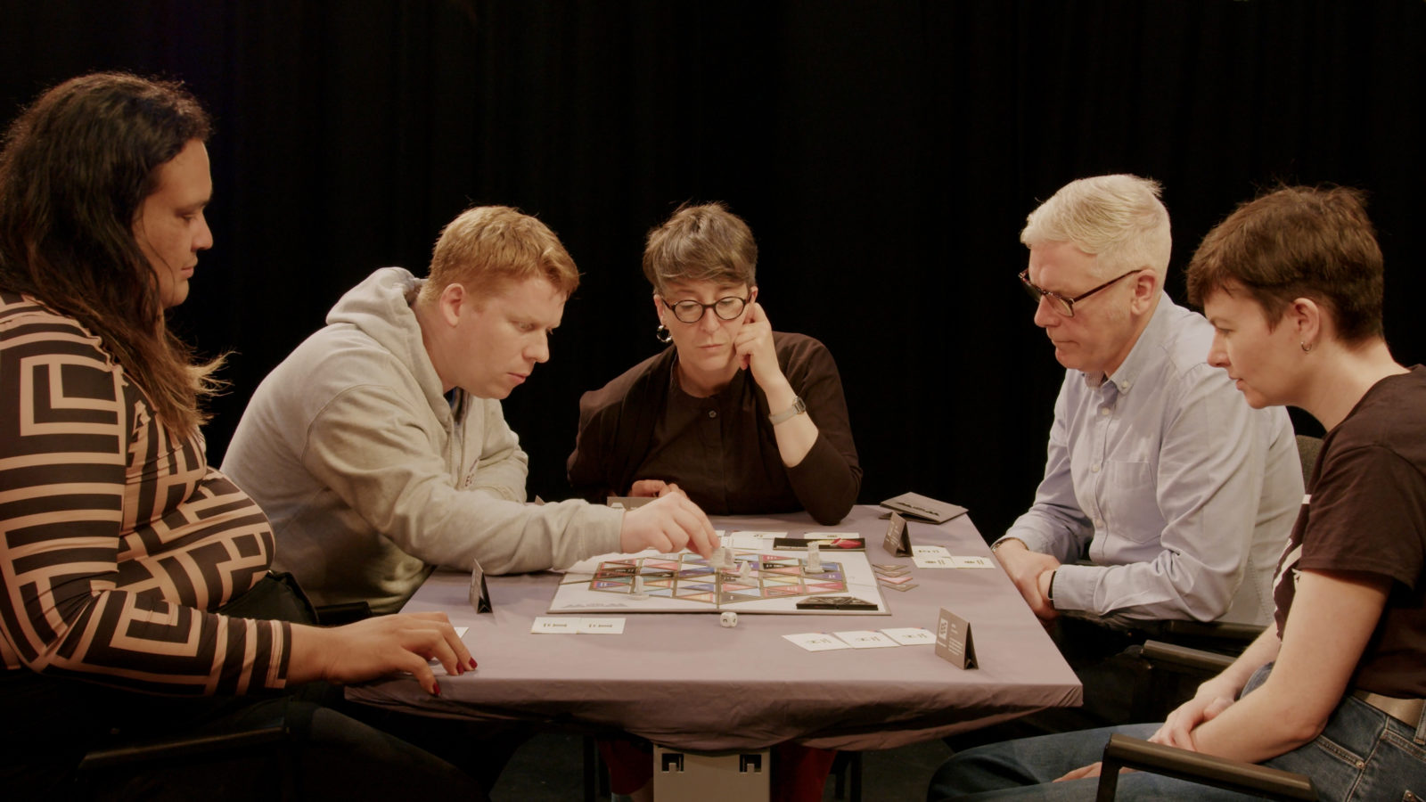 5 people sit around the table playing the game, they are all looking thoughtful and concentrating, one person reaches out and moves a piece on the board.