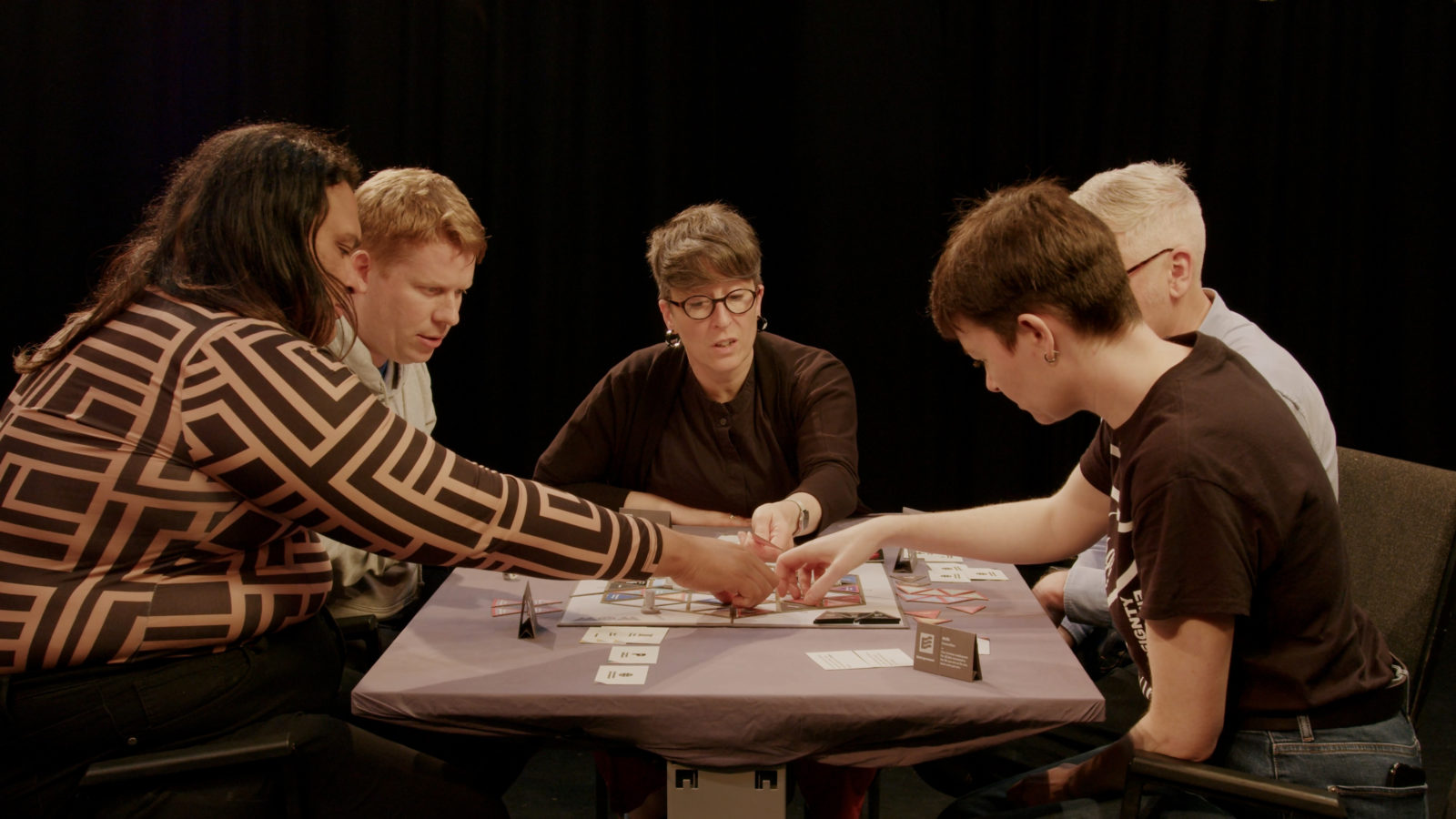 5 people are photographed around the table playing the board game. 3 people reach for things on the board and all of them are looking thoughtful.