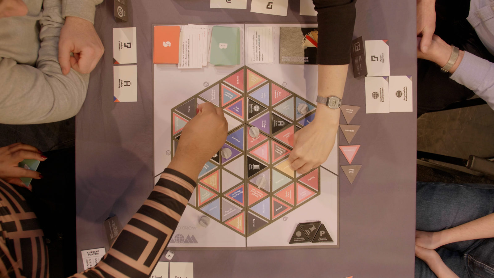 The board game is photographed from a Birds Eye view. 2 hands from opposite ends of the table reach for cards from the board.