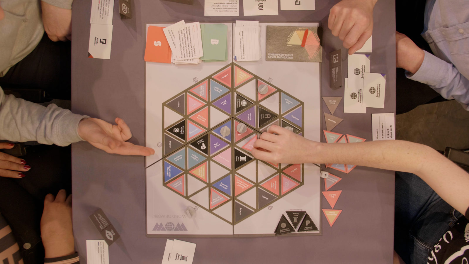 A Birds Eye view shot of the board game being played, one hand picks up a card and another points at the board from the other side of the table. The cards are small triangles in different colours that make up a hexagon in the middle of the board.