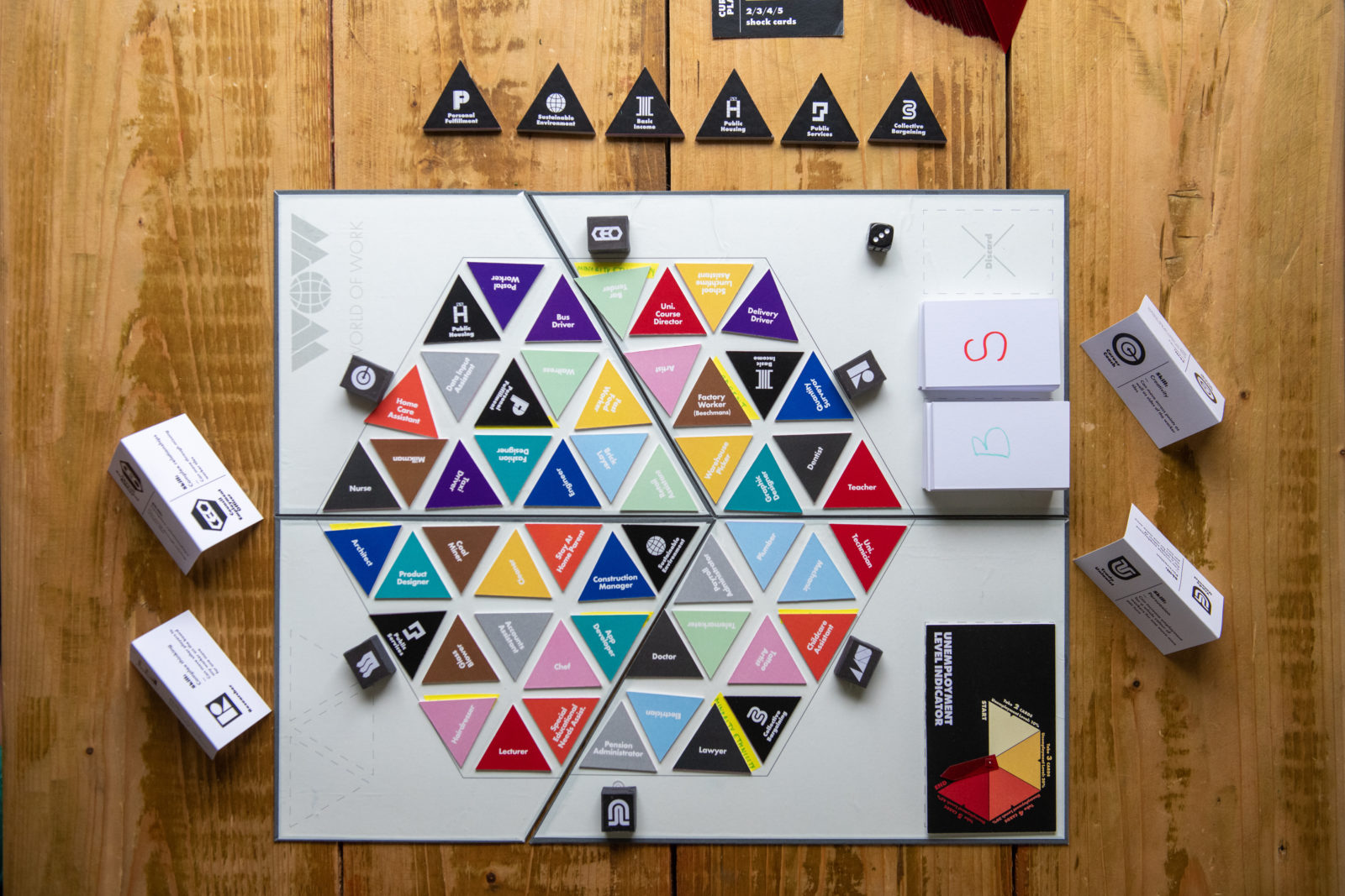 A board game is set up on a wooden table. Colourful triangular counters are set up on the board, black counters on the table around it and black and white cards.