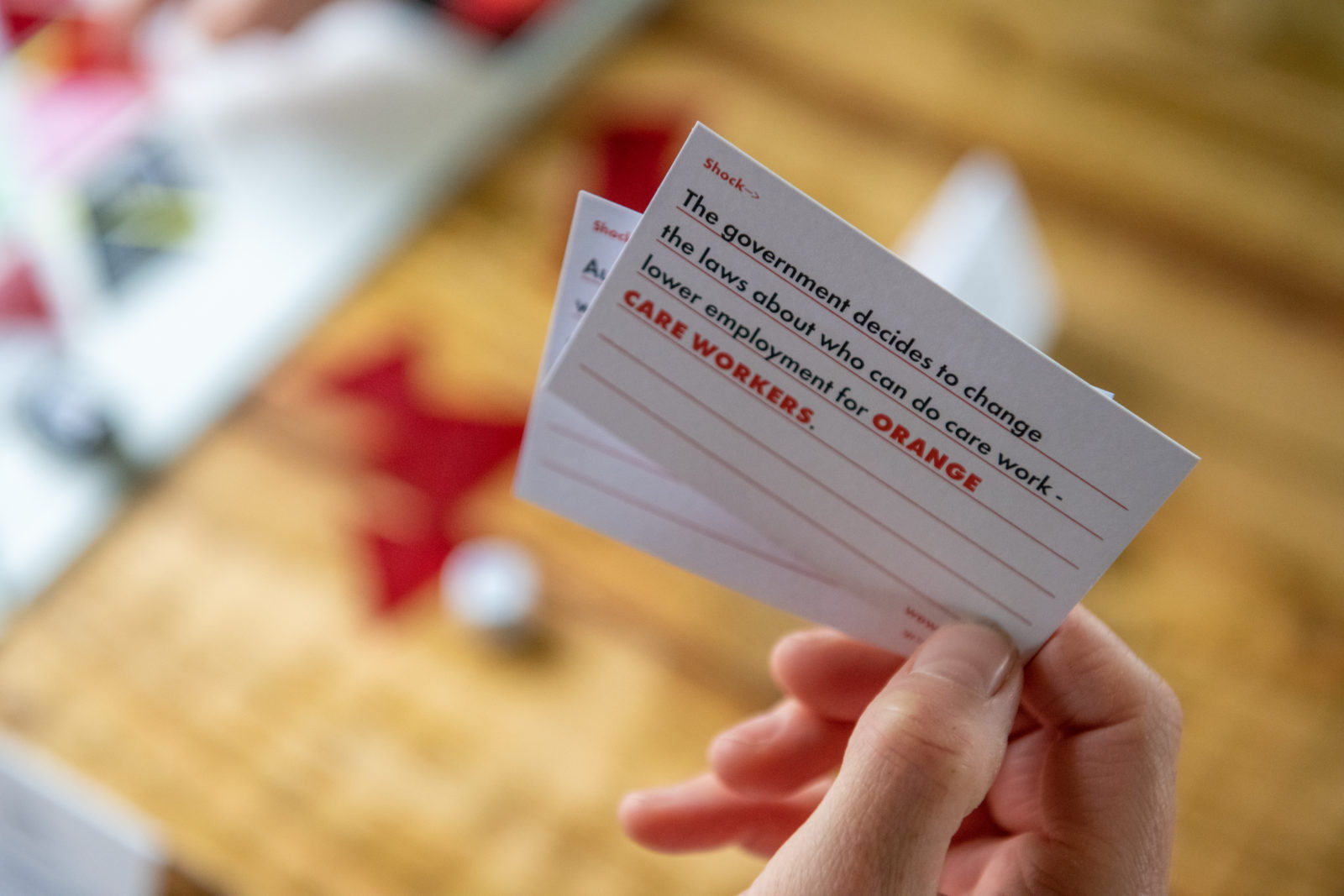 A hand hold a small white card with lines on it. In black and orange text the card reads 'The government decides to change the laws about who can do care work - lower employment for Orange Care Worker'.