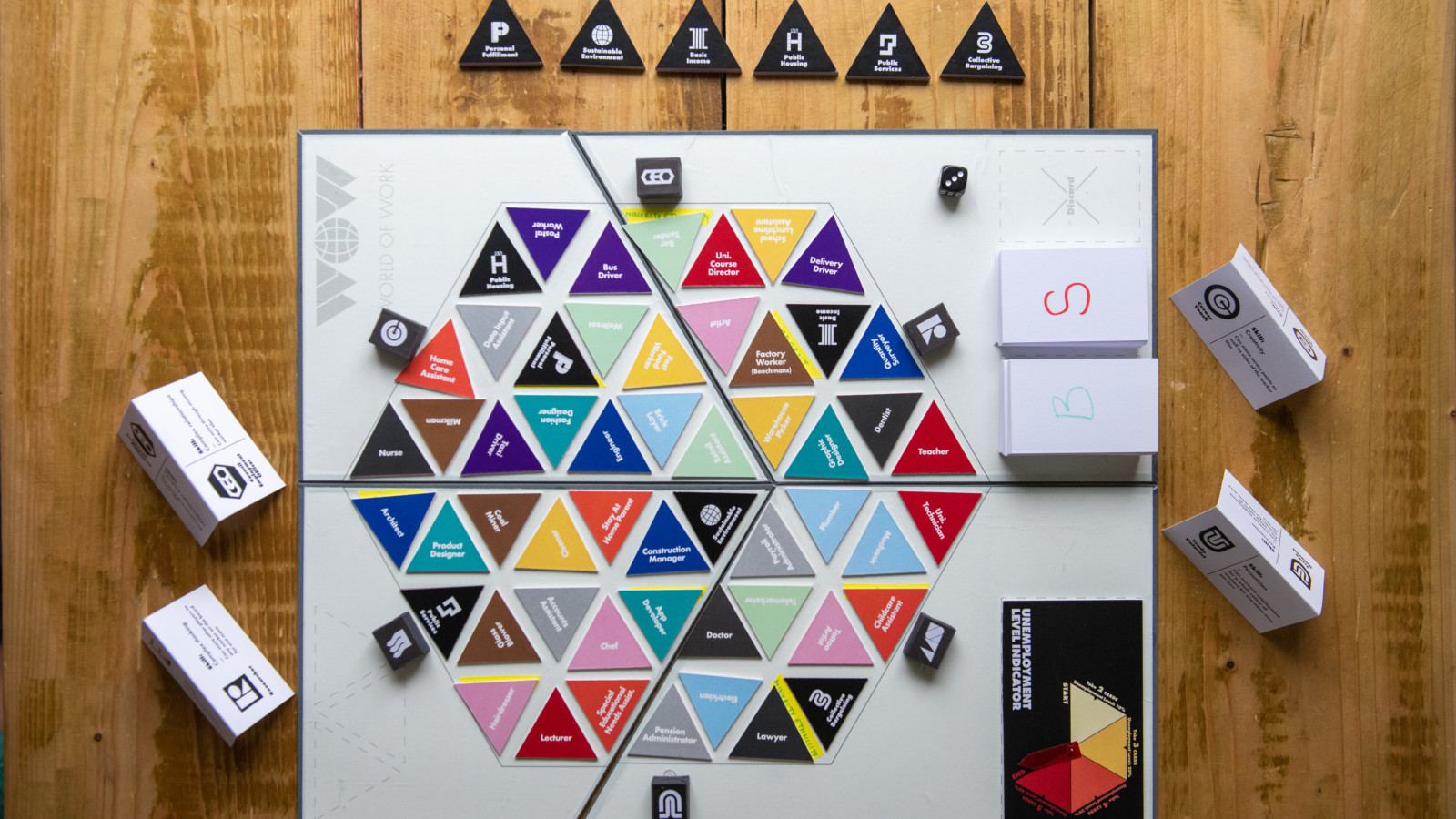 A board game is set up on a wooden table. Colourful triangular counters are set up on the board, black counters on the table around it and black and white cards.