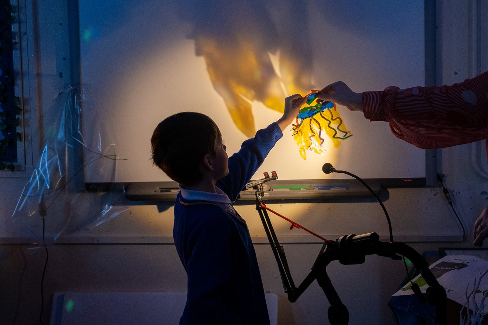 In a darkened room a school child holds up a yellow piece of plastic in the shape of an octopus in front of a bright light, projecting the colours and shadows on to a white screen. An adults arm reaches in from off camera to help.