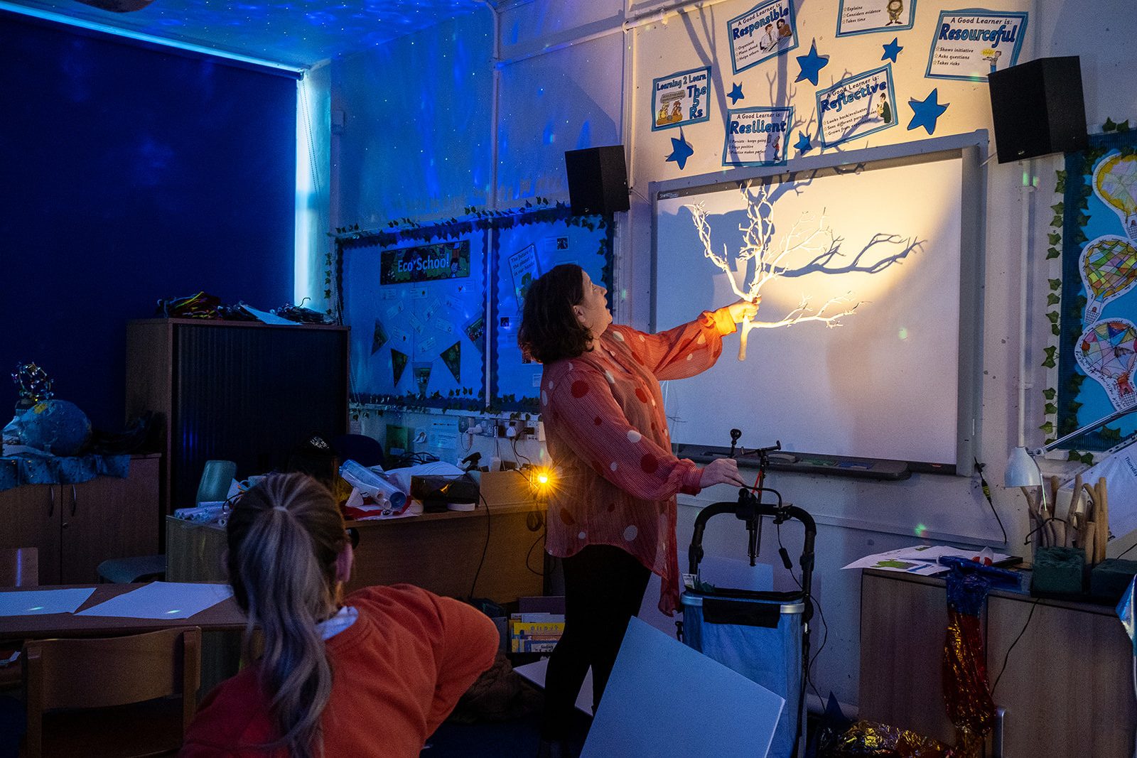 In a darkened classroom artist Cathy Cross holds up a tree like structure in front of a bright light, illuminating the beautiful structure and projecting it's shape on to the wall. In the background a projector is shining stars and lights on to the wall.