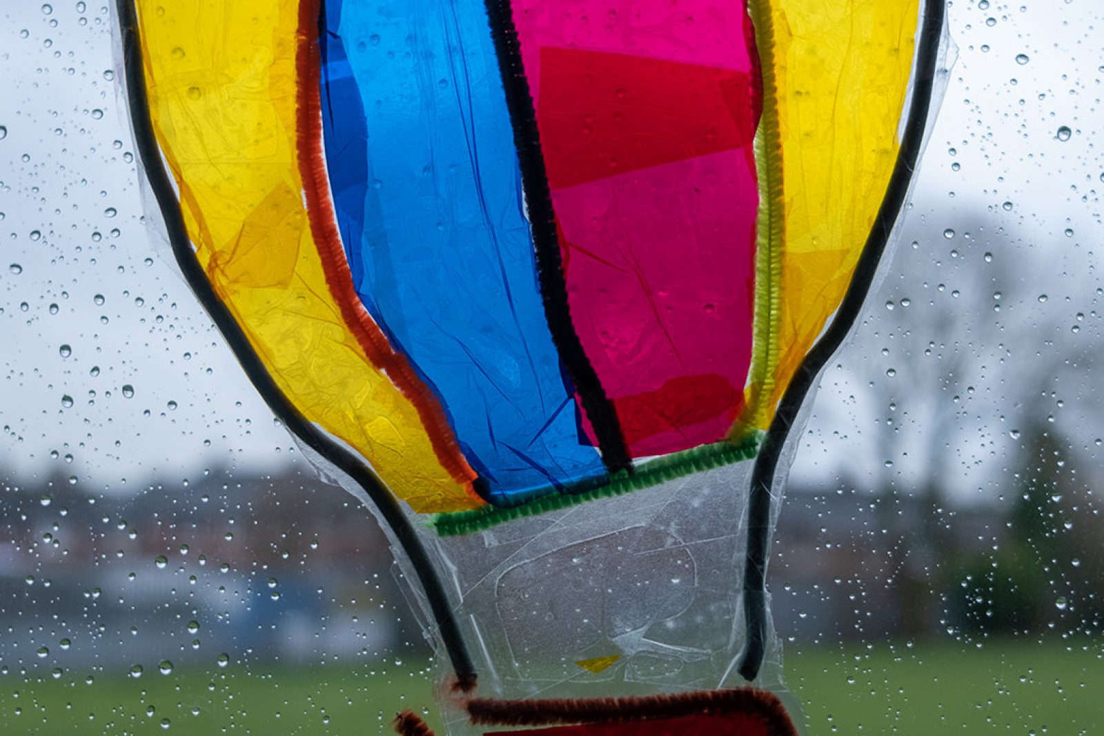 A colourful cut out of a hot air balloon made of clear plastic is stuck to a window letting the light shine through it. The bright colours contract against a grey and rainy day outside the window.