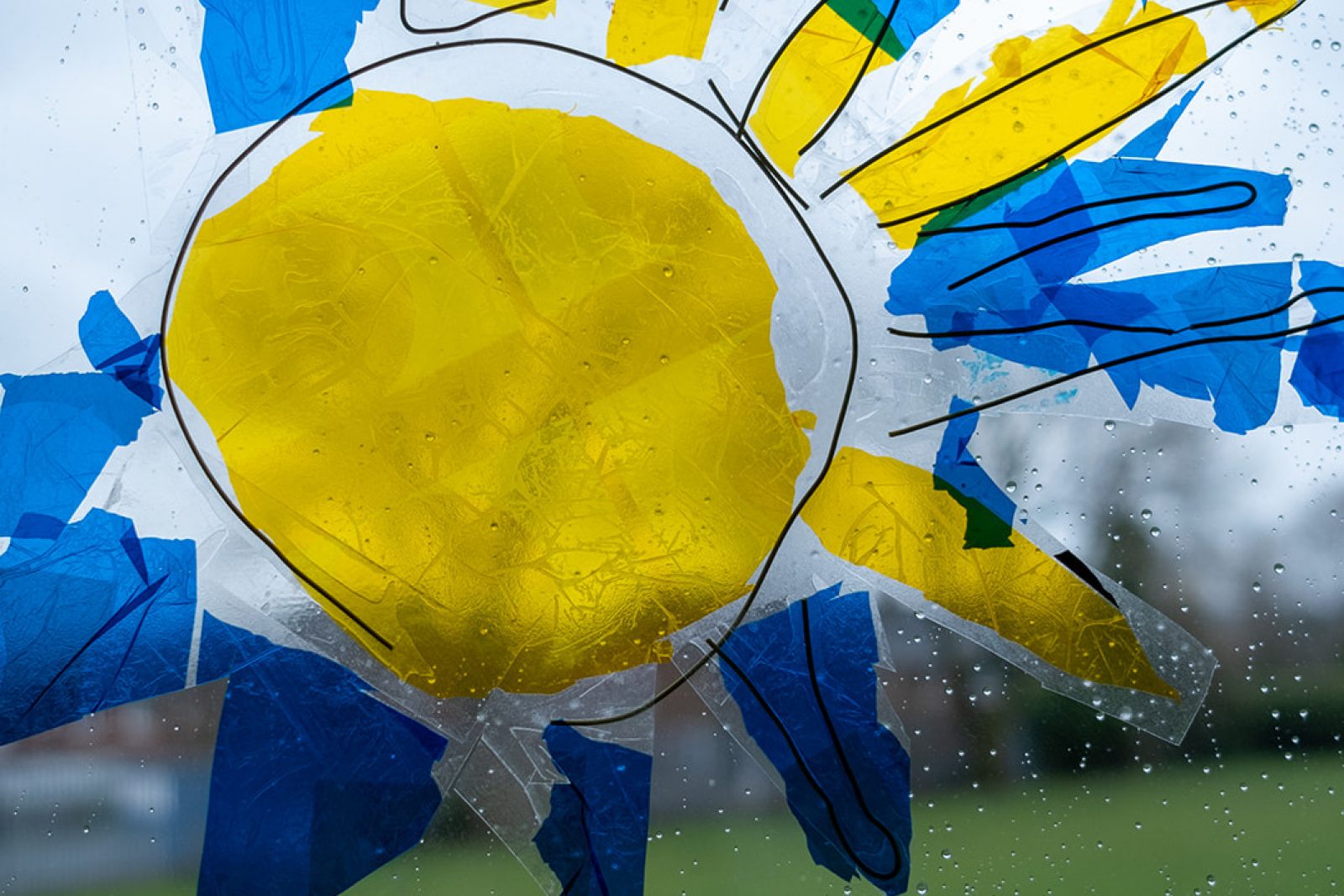 A colourful cut out of a sun made of clear plastic is stuck to a window letting the light shine through it. The bright colours contract against a grey and rainy day outside the window.