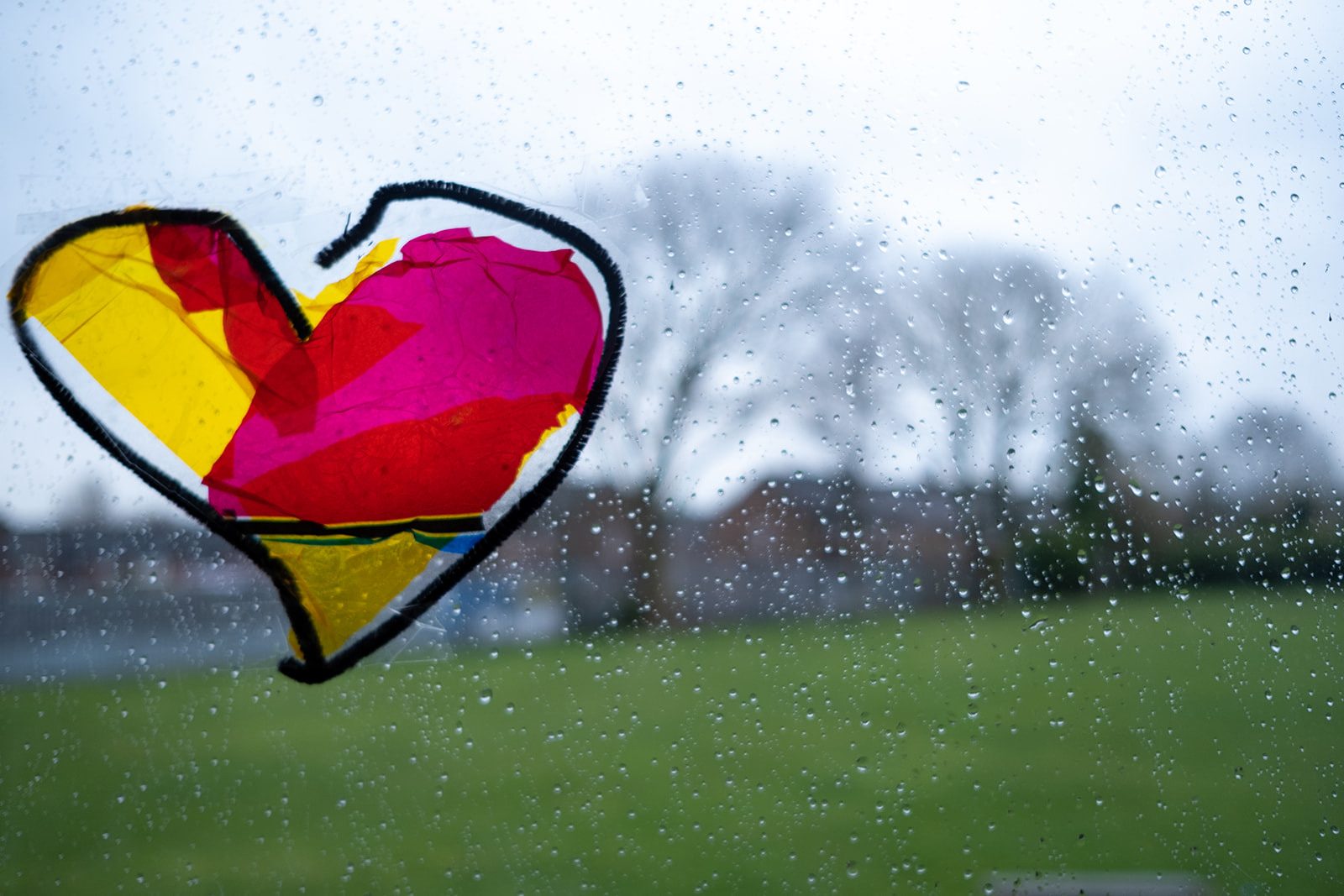 A colourful cut out of a heart made of clear plastic is stuck to a window letting the light shine through it. The bright colours contract against a grey and rainy day outside the window.