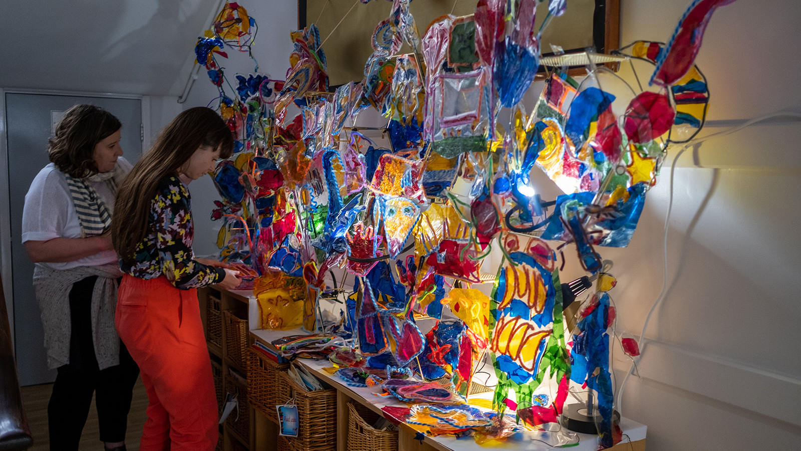 Artists Billy Colours and Cathy Cross stand in front of an incredibly bright and colourful sculpture made of different coloured transparent plastics. The sculpture sits on a long shelf and is illuminated from behind making the colours shine brightly.