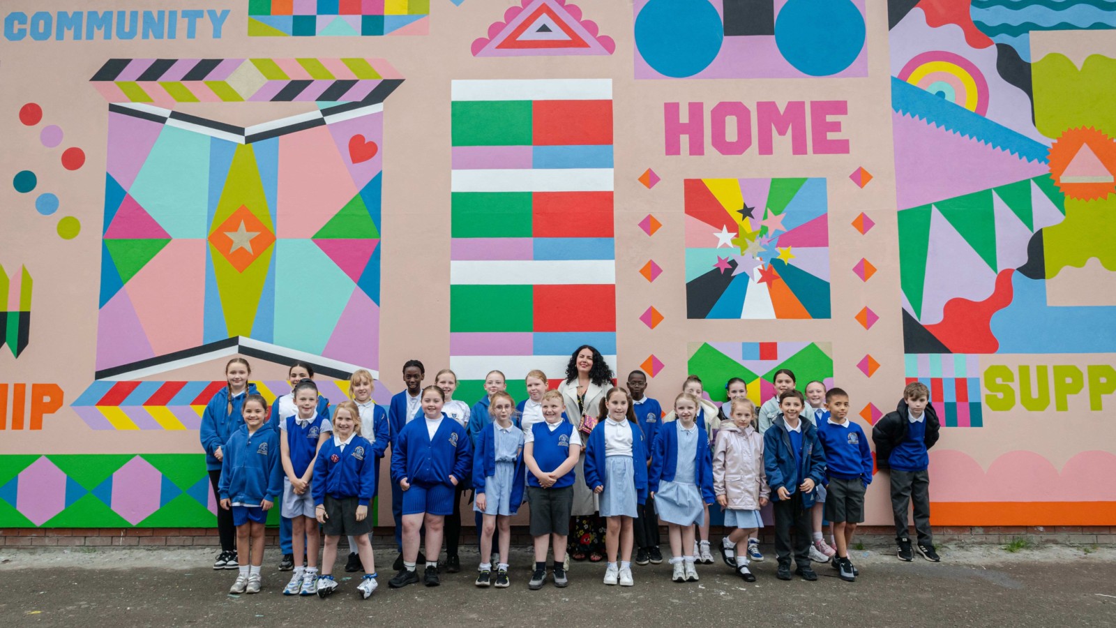 The schoolchildren stand in front of the mural with Cherie smiling at the camera. The mural is made up of vibrant, geometric shapes and patterns, with words such as ‘Home’, ‘Friendship’ and ‘Community’ on it.