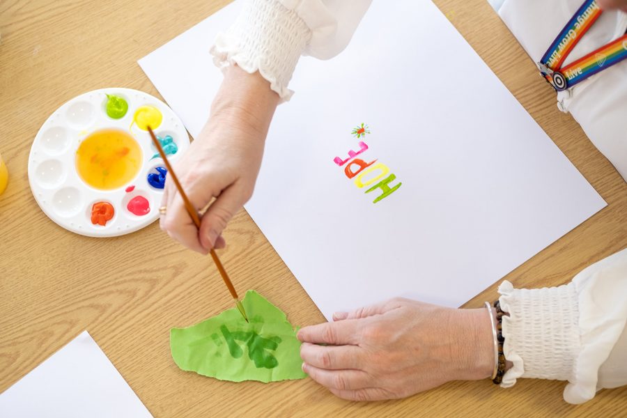 A paint palette is filled with multicoloured paints on top of a table. Someone is sat at the table working on a white piece of paper. On the paper the word hope is painted in green, yellow, orange and pink paint.