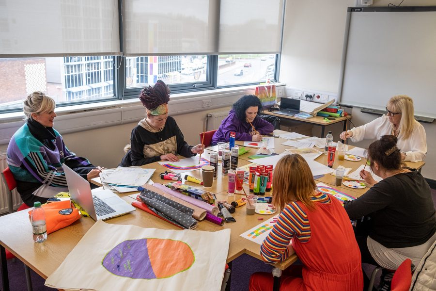 A group of women are sat around a table covered in art materials; paints, pencils, colourful paper and big white sheets. The women are all absorbed in painting on their own sheet and conversation together.