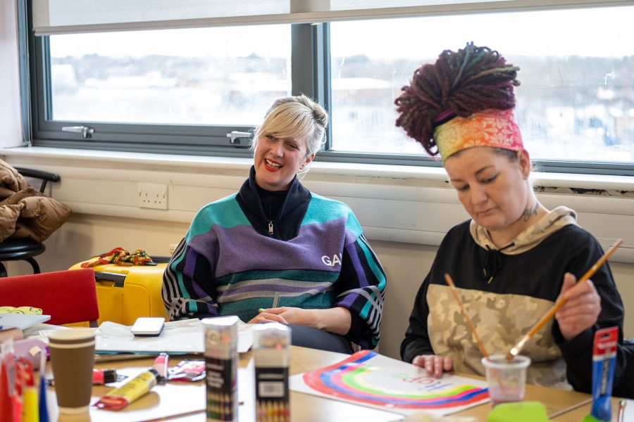 Annie Nicholson sits at a table with a big smile on her face. Next to her is a person wearing a colourful headband painting a rainbow on a piece of white paper. On the table are tubes of paint, coloured pencils, paper and other craft materials.