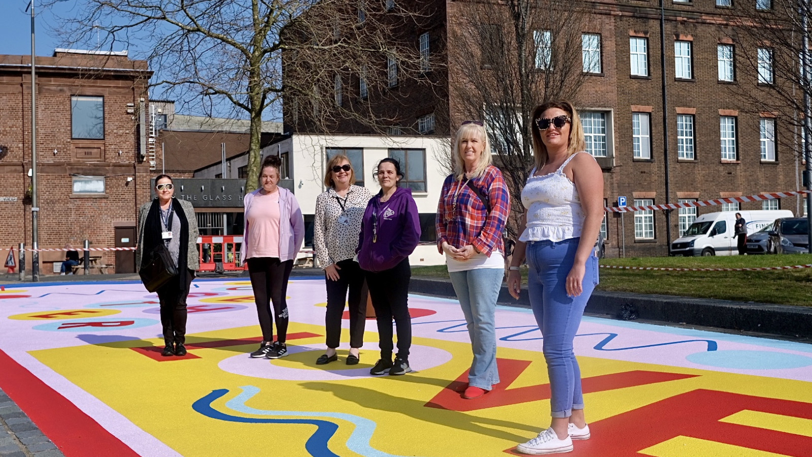 A group of women of different ages stand together on a bold and bright floor mural. You can read the word 'love' under their feet.