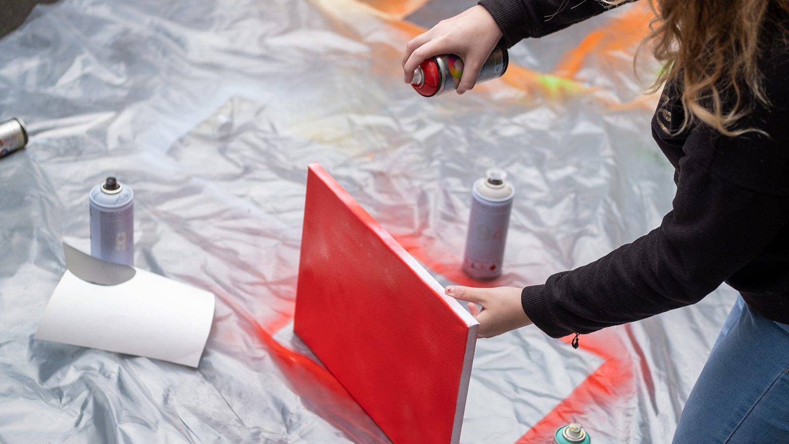 On a large sheet of plastic someone spray paints a white canvas with bright red spray paint.