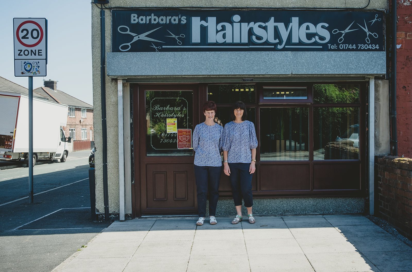 Two people stand smiling in matching outfits in front of a wooden panelled shop window. Above them is a large sign with a silver design on a black background which reads 'Barbara's Hairstyles' with scissors.