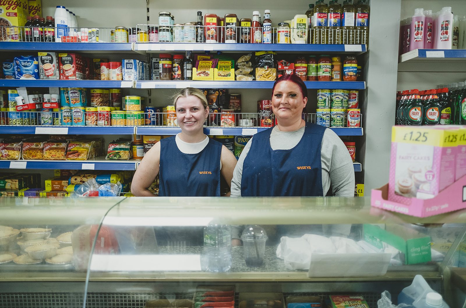 Two people stand behind a shop counter smiling. They each wear a tabard apron with the word 'Wiseys' and behind them are some shop shelves full of tins and packets of food.