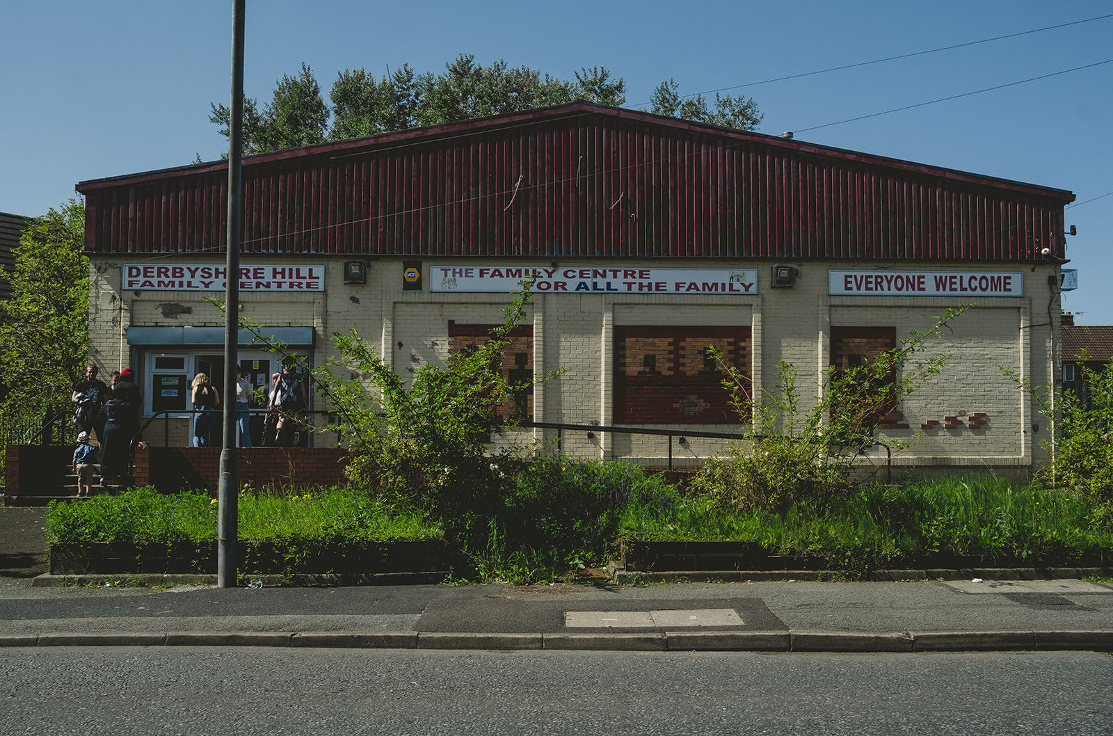 Derbyshire Hill Family Centre. A single story building painted white with a red, corrugated metal roof. Signs on the building say "A Family Centre For All The Family" and "Everyone Welcome"