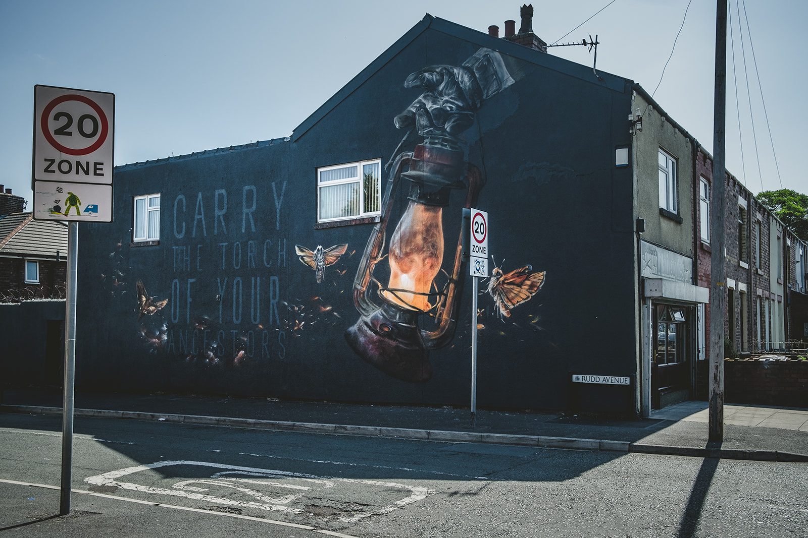 We Follow The Light mural by Nomad Clan. A large wall is painted with a mural depicting a gloved hand holding an old industrial lamp. The lamp glows orange and attracts moths towards it. To the side of this image are the words 'Carry the torch of your ancestors'