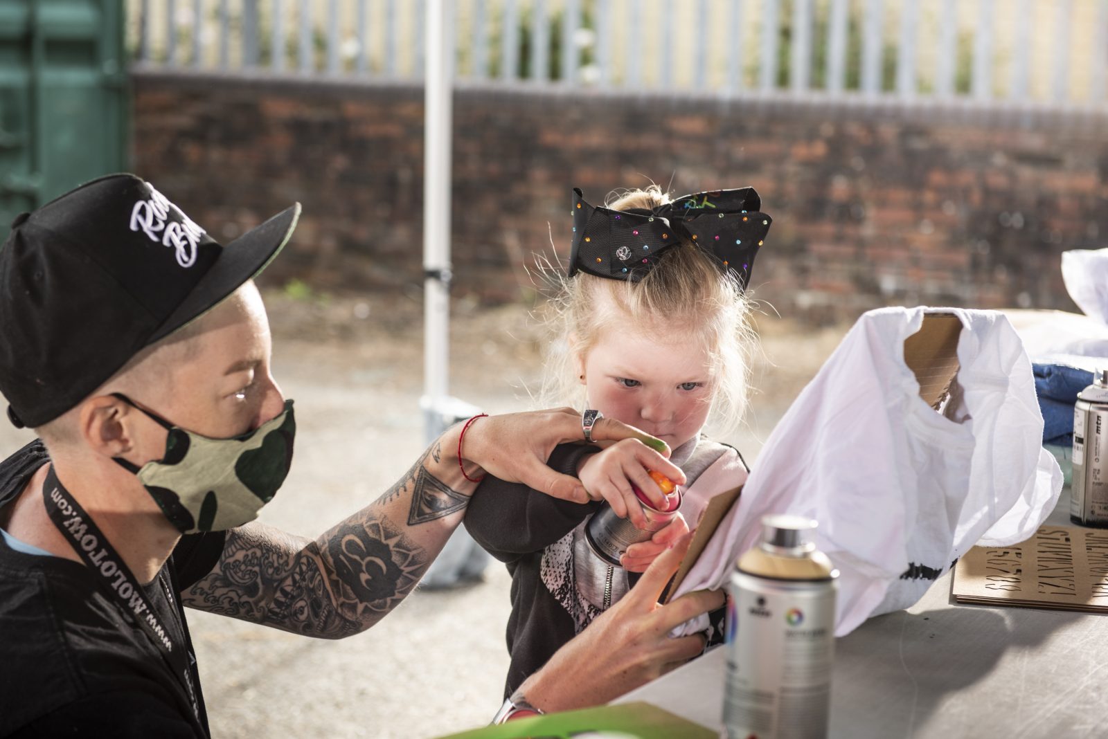 Nomad Clan's Jay Gilleard helps a little girl spray paint a stencil design onto a t-shirt. Jay wears a black baseball cap, and a black t-shirt, they also have a khaki green and black face mask. The little girl is also dressed in black and has a large bow in her hair.