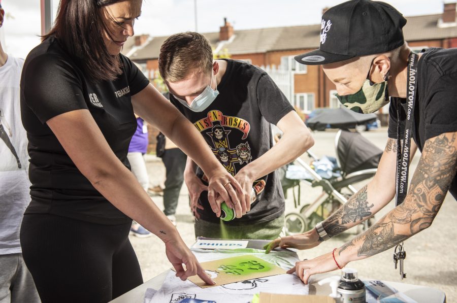 Nomad Clan's Jay stands holding a stencil down on some paper while two people spray green spray paint over the stencil to create a design.