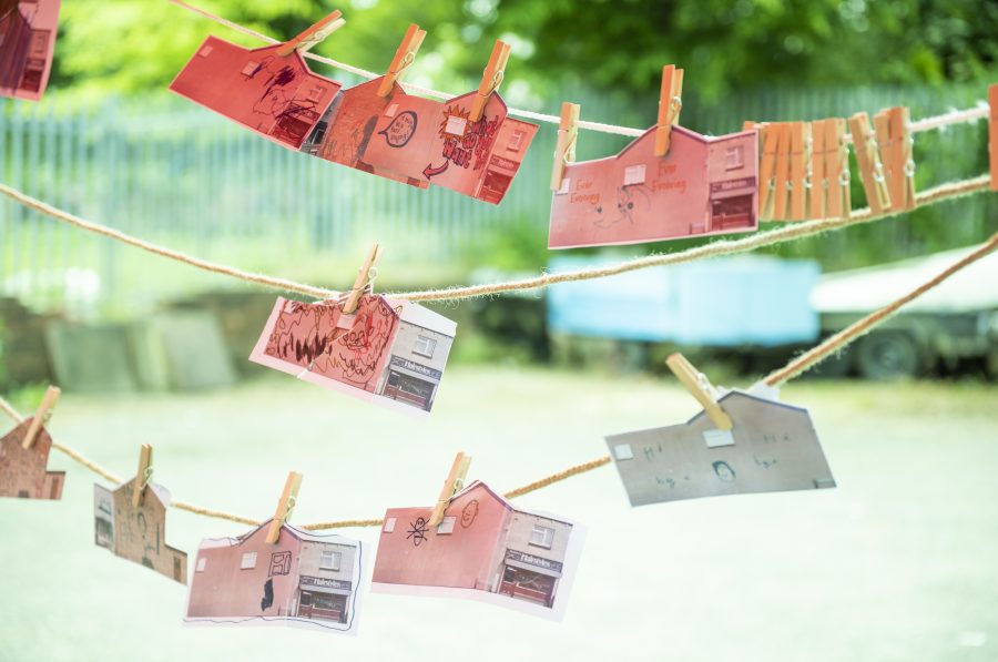 Lots of paper cut outs in the shape of a building are pegged on three washing lines. On each cut out are drawings, words and designs.