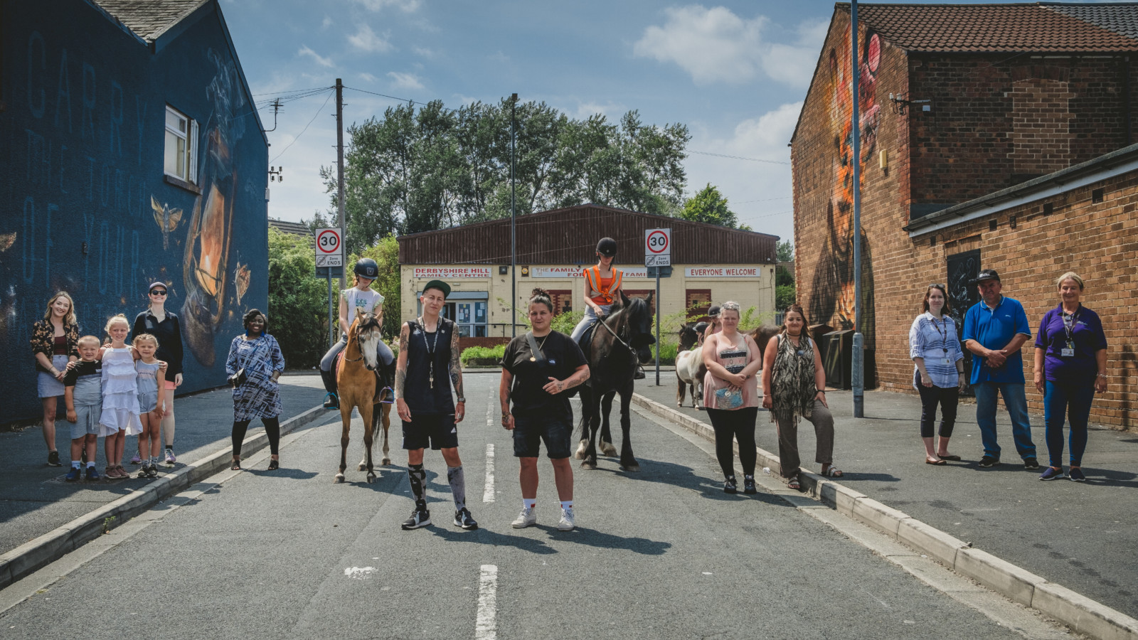 On a sunny day a groups of people stand on a street in smaller, socially distanced groups facing the camera. Behind them, on either side of the street are two buildings each with a large mural on the wall.