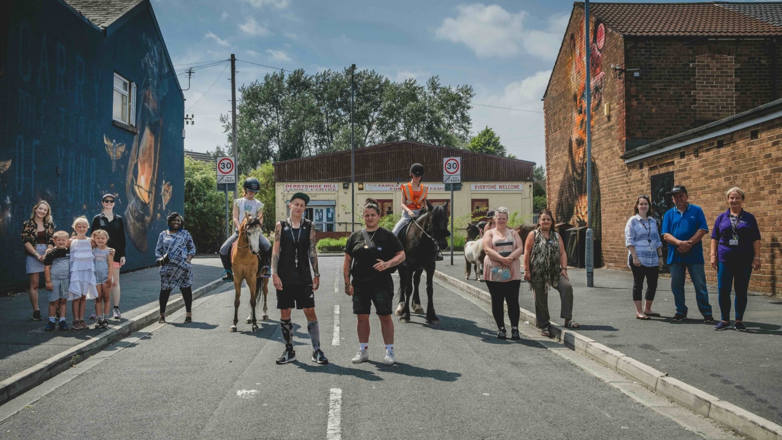 Several people stand in the road between the two murals looking at the camera, it is a sunny day.