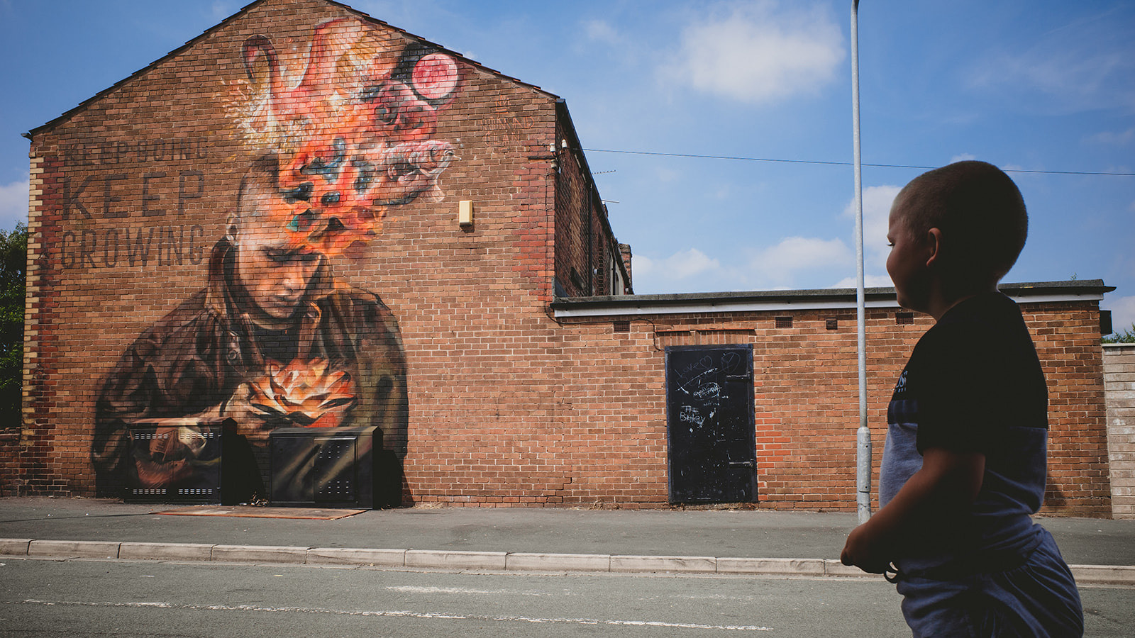 A child stands silhouetted buy a bright sunny day, they are looking at a building across the road and on the red brick wall there is a large mural painting of a person with a coloured burst rising from their head.