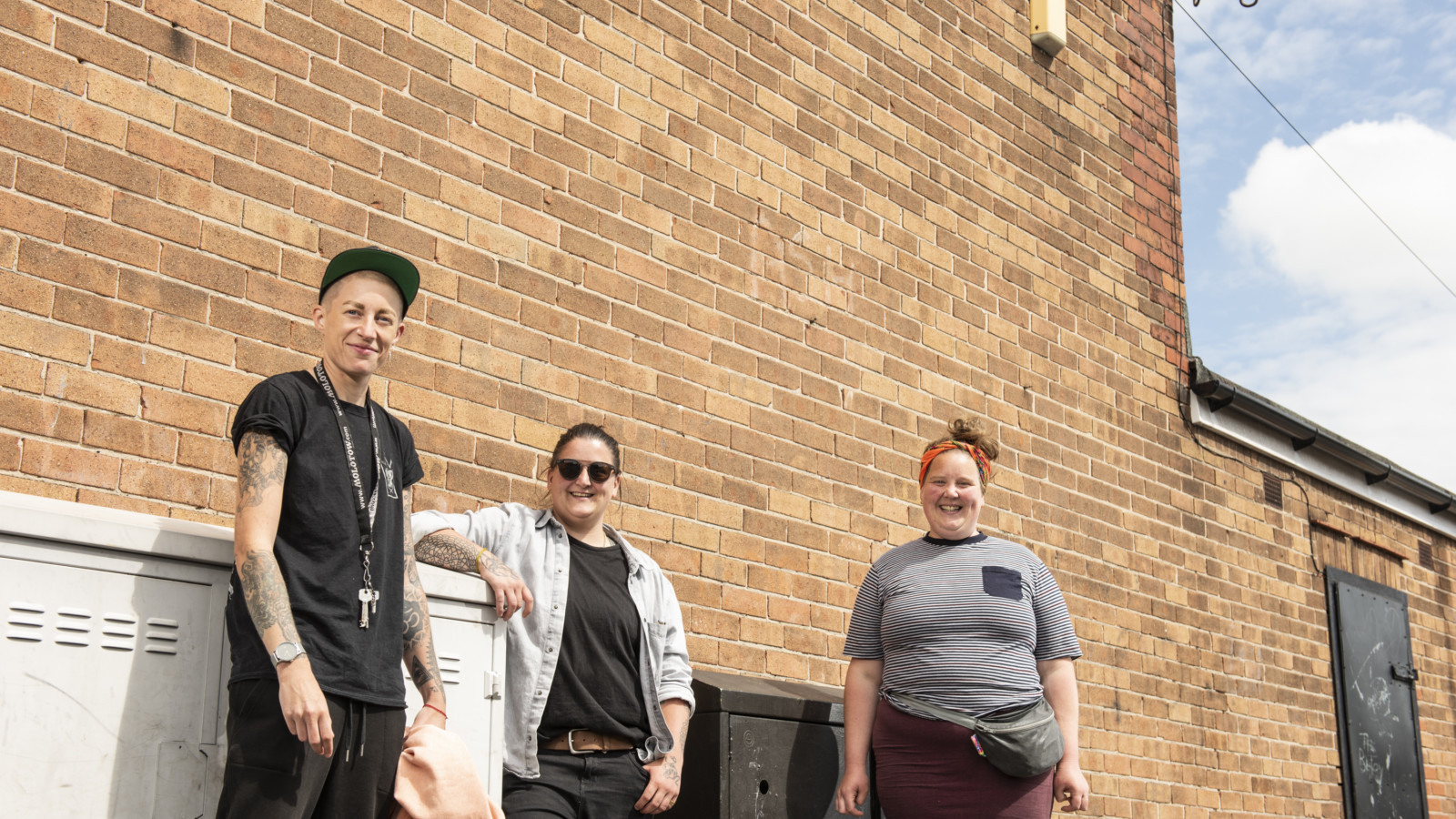 Nomad Clan and Michelle Wren stand in front of a large brick wall in the bright sunshine. They are all smiling.