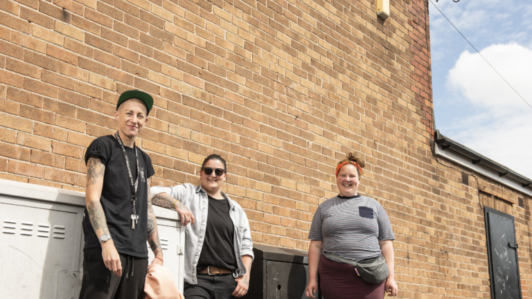 Nomad Clan and Michelle Wren stand in front of a large brick wall in the bright sunshine. They are all smiling.