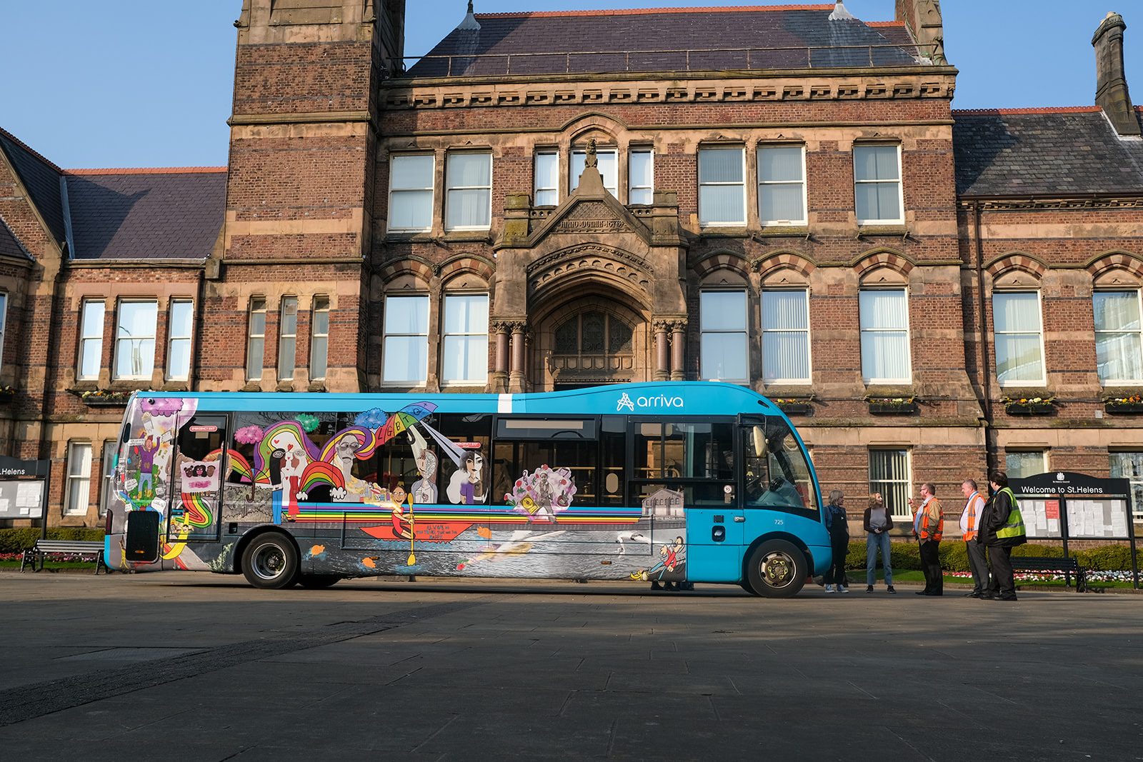 An Arriva North West bus is parked in front of St Helens Town Hall. The bus is decorated with colourful illustrated designs of rainbows, people and a big rainbow coloured umbrella.