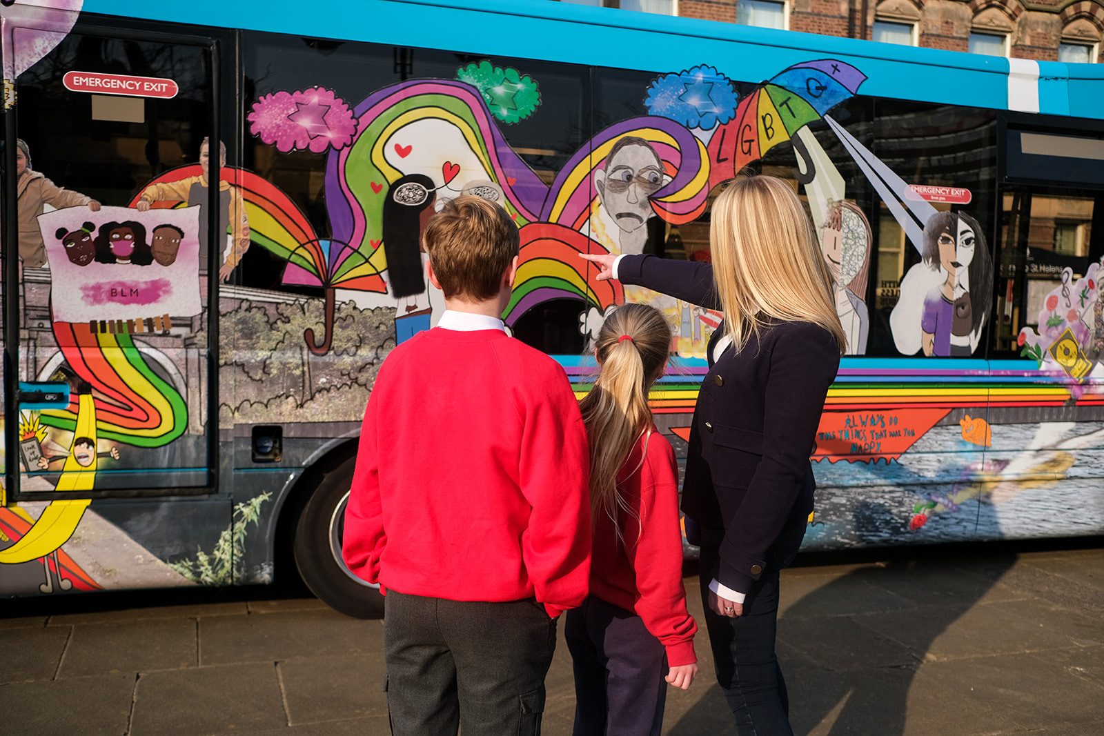 Three people, two children in school uniform and one adult, stand looking at the side of the decorated bus.