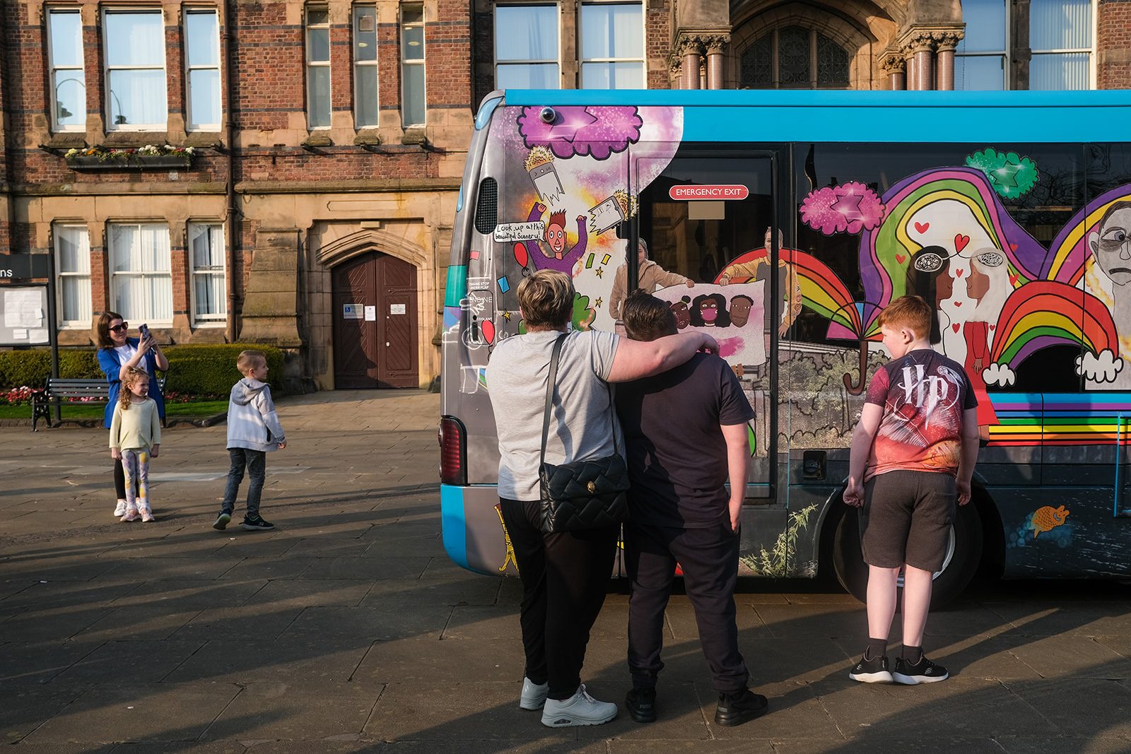 Children and adults stand around a colourful decorated Arriva North West bus. Two people stand one with their arm around the other, looking at the designs on the bus.