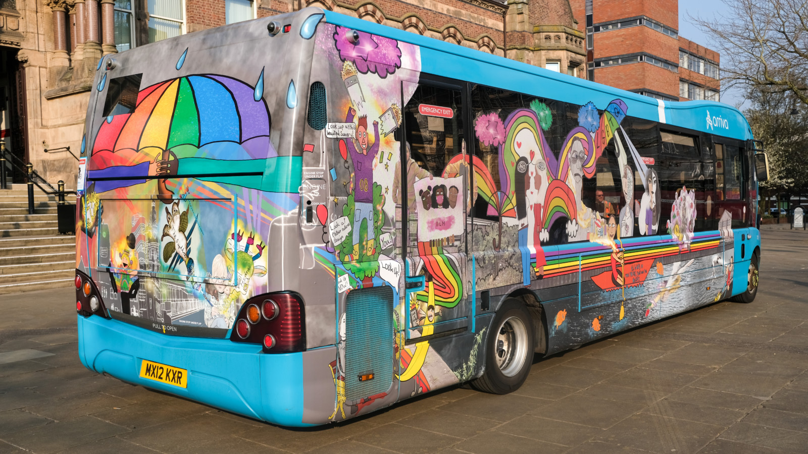 An Arriva bus is parked in front of St Helens Town Hall. The bus is decorated with brightly coloured illustrations and designs. The rear of the bus has a large rainbow coloured umbrella.