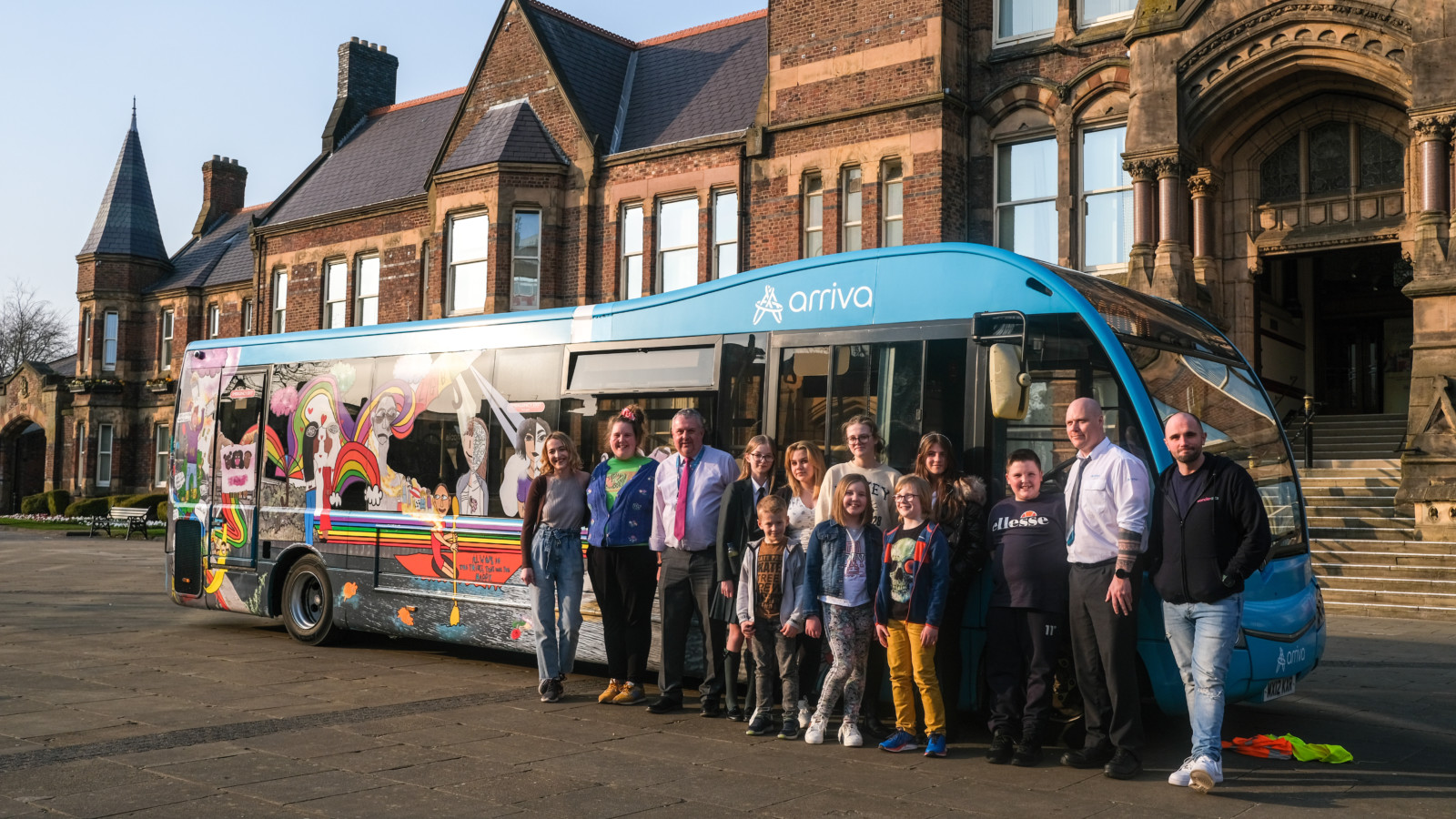 A group of people stand at the front end of an Arriva bus in front of St Helens town hall. The bus is decorated with brightly coloured illustrations. The evening light illuminates the bus.