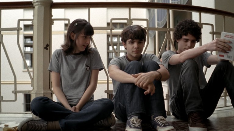 Film still featuring three teenagers sitting on the floor, underneath school chairs and tables