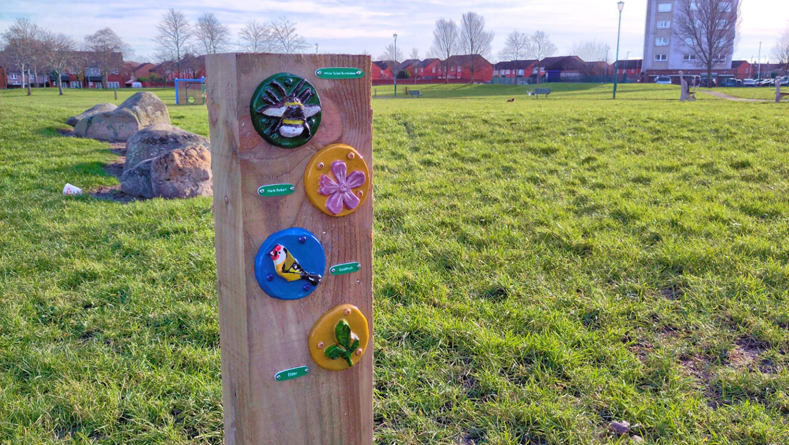 Photograph of four ceramic tiles featuring leaves, birds and bugs The tiles are attached to a wooden post, outdoors