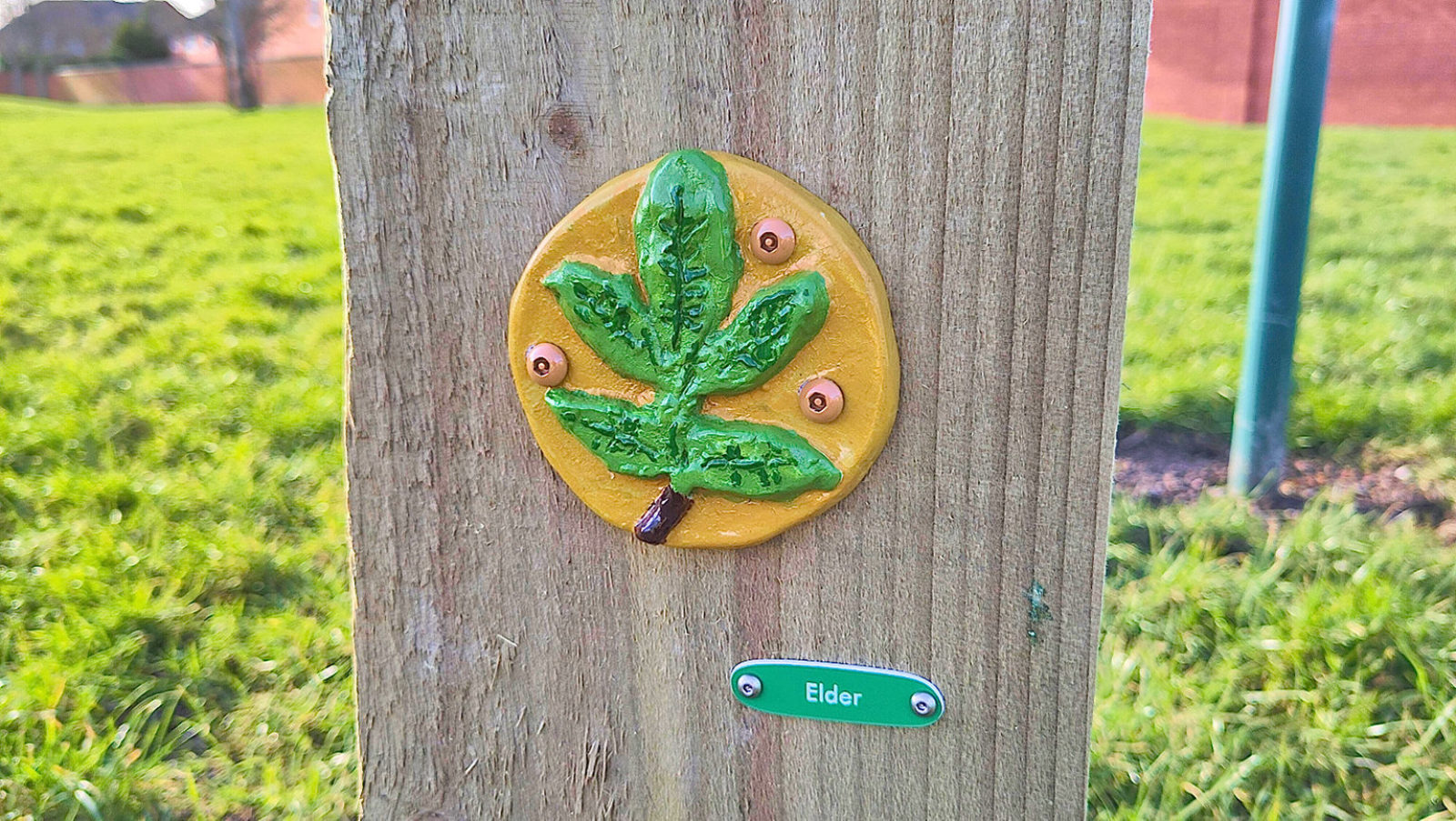 Photograph of a ceramic tile featuring a hand painted leaf. The tile is attached to a wooden post, outdoors