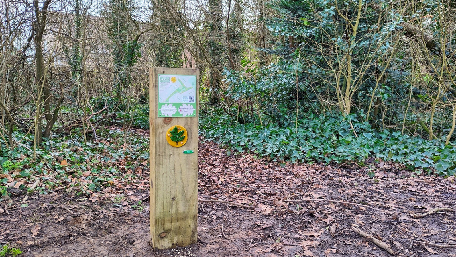 Photograph of a wooden post in a park, with a map and a small artwork in the shape of a leaf attached to it