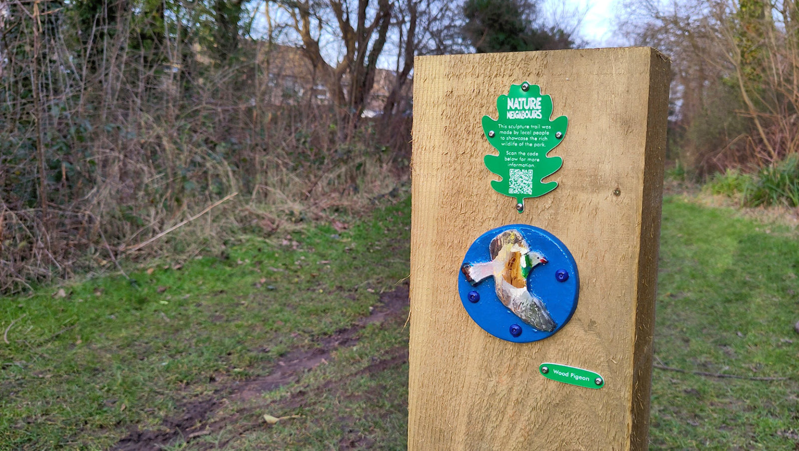 Photograph of a wooden post in a park. Attached to the post is a small plaque and a small handmade artwork of a bird