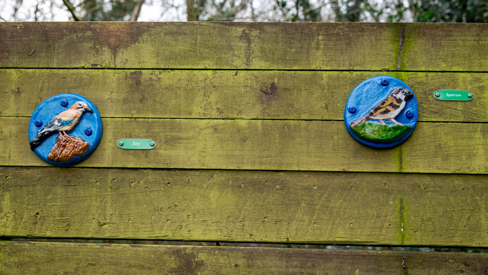 Photograph of a fence with two small hand-painted tiles featuring birds attached