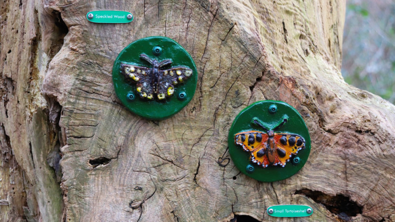 Photograph of two ceramic tiles featuring butterflies, attached to a tree stump. The tiles are handmade and hand painted