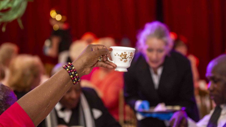 A black person wearing a red shirt and shiny bracelet holds up a posh tea cup at a posh afternoon tea event
