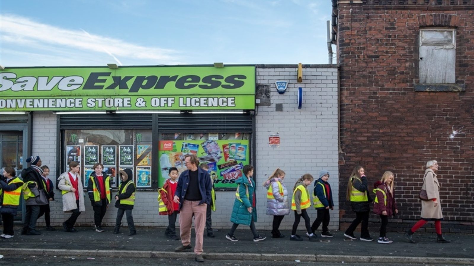 A group of children wearing coats and yellow hi vis jackets walk on a pavement, single file in a line past a shop called Save Express. They are lead by a woman in a beigt coat. A man walks across the road towards the camera, looking out for traffic.