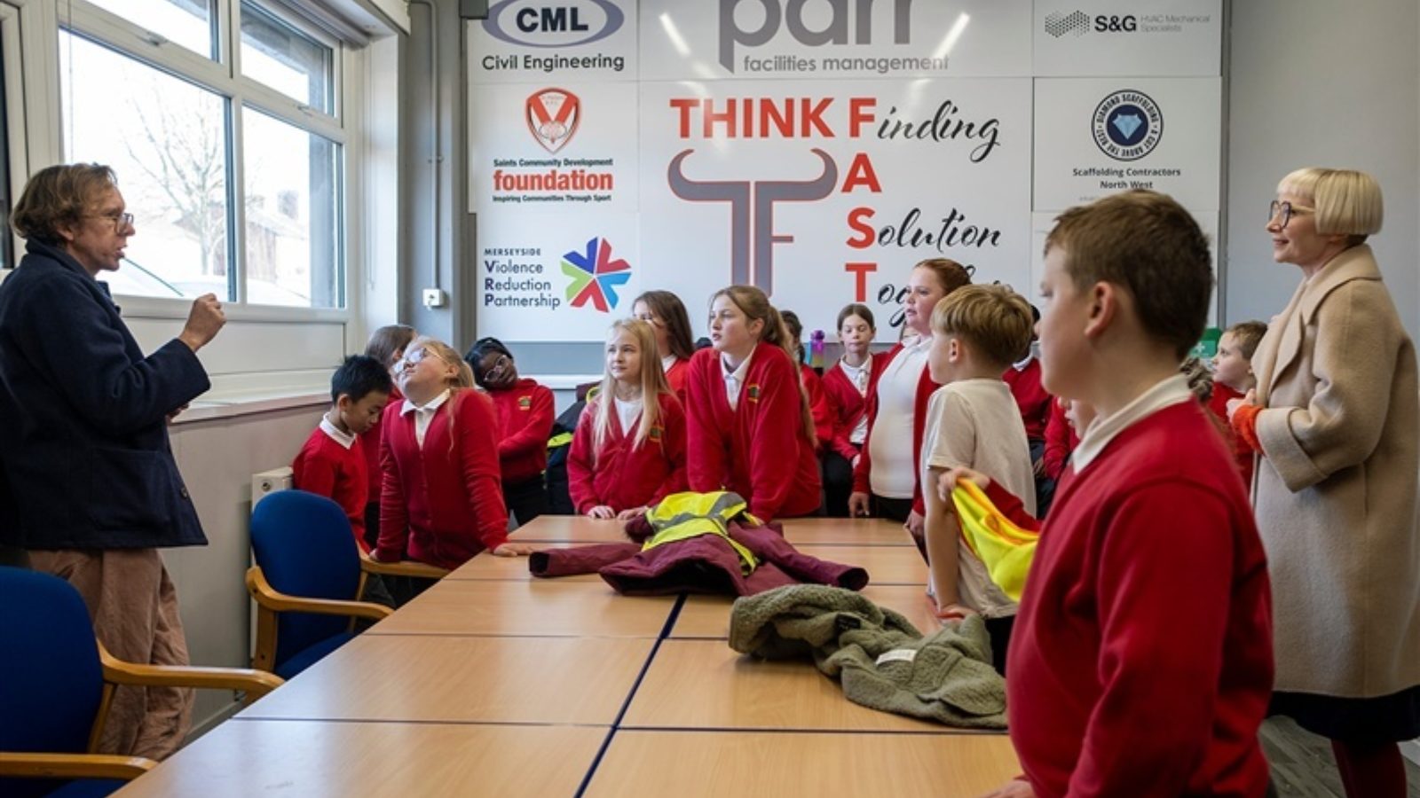 A group of children in stand gathered around a large table watching and listening to a man who is standing in front of a window talking. A woman in a beige coat stands behind them, also listening.