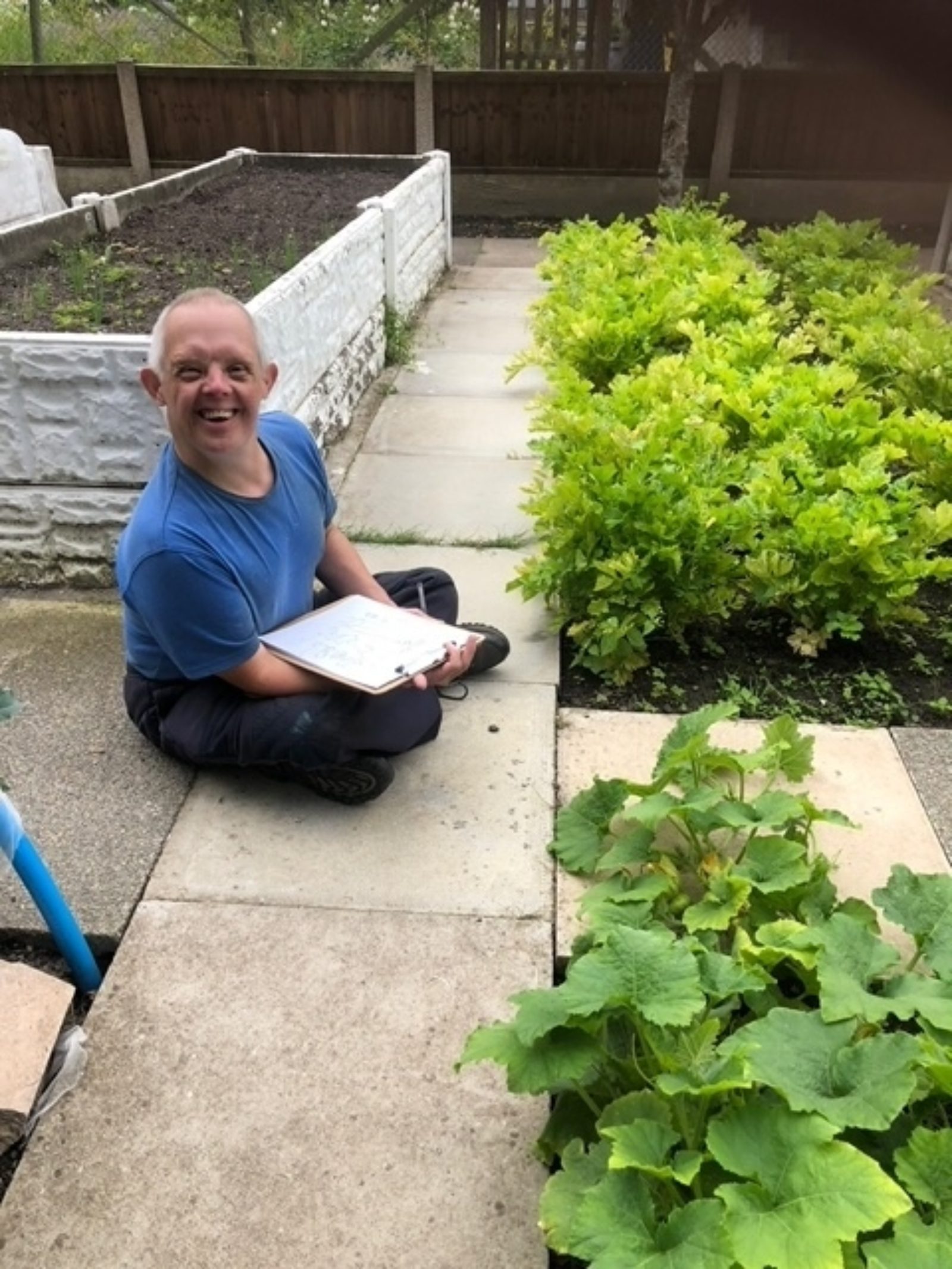 A workshop participant from Buzz Hub St Helens is smiling at the camera, sat on the floor in front of some green leafy plants with a pencil and clipboard.