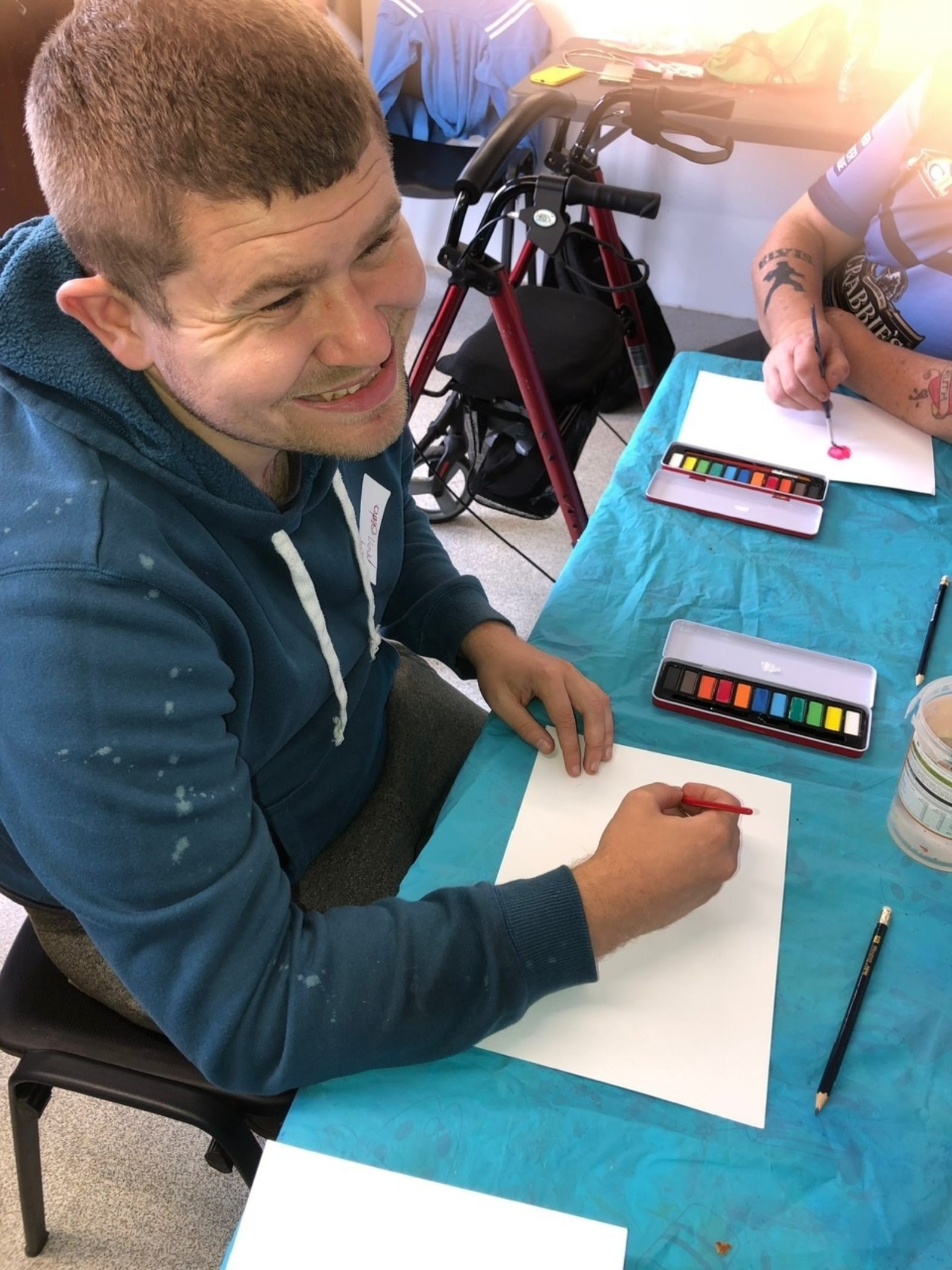 A man smiles broadly as he is photographed drawing at a table, there are watercolour palettes on the table.