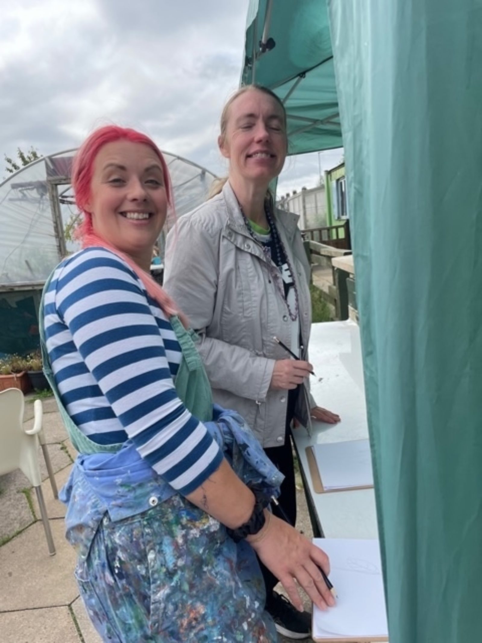 Laura Kate and a member of Buzz Hub St Helens smile for the camera at Incredible Edible community garden with pencils in their hands and paper in front of them.