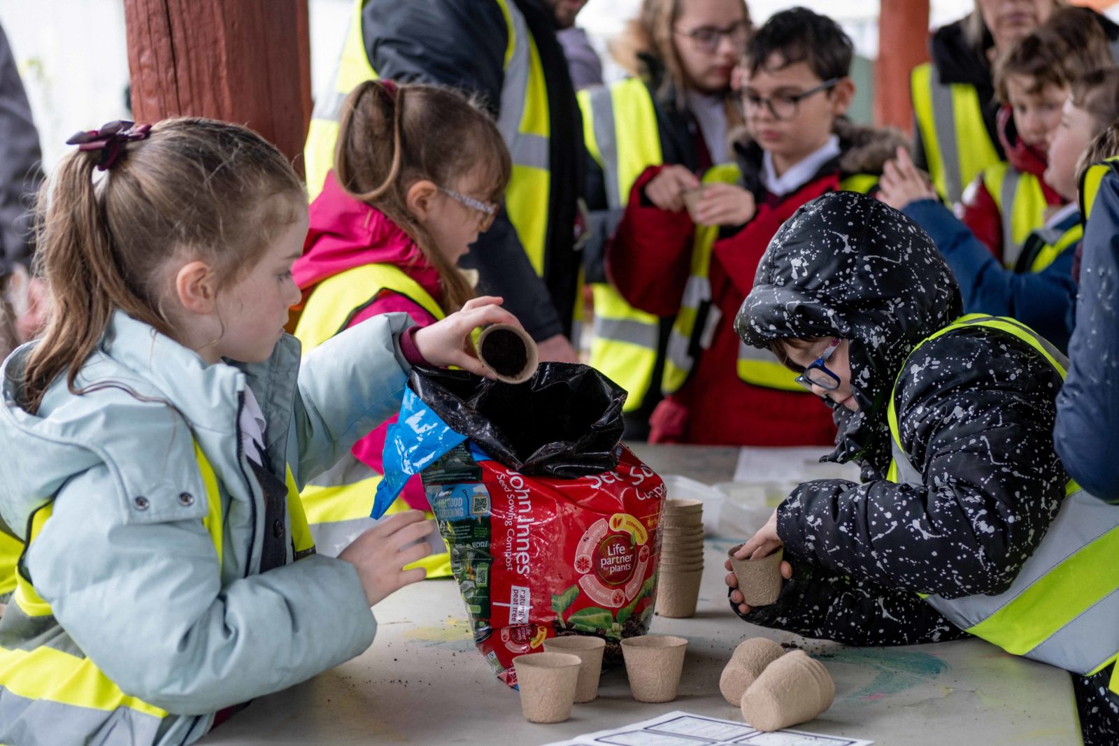 Schoolchildren gather around a table, filling plant pots with soil.